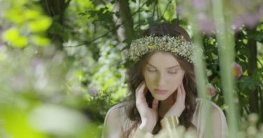 A young woman with a wreath of flowers on her head stands in a white light dress in the garden among green leaves and flowering plants. slow motion