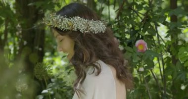 A young woman with a wreath of flowers on her head stands in a white light dress in the garden among green leaves and flowering plants. slow motion