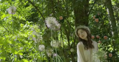 A young woman with a wreath of flowers on her head stands in a white light dress in the garden among green leaves and flowering plants. slow motion