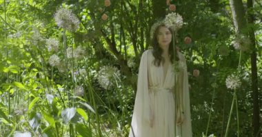 A young woman with a wreath of flowers on her head stands in a white light dress in the garden among green leaves and flowering plants. slow motion