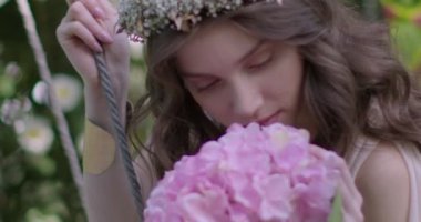 A young beautiful girl in a wreath of flowers swings on a swing among a green garden. Slow motion