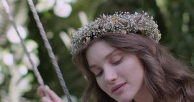 A young beautiful girl in a wreath of flowers swings on a swing among a green garden. Slow motion