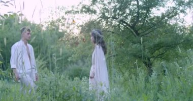 A young couple in white light robes walk towards each other through a spring flowering garden. Slow motion