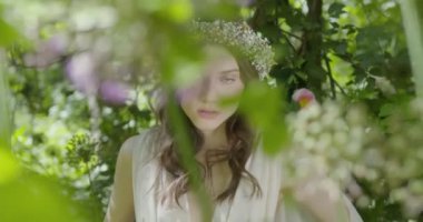 A young woman with a wreath of flowers on her head stands in a white light dress in the garden among green leaves and flowering plants. slow motion