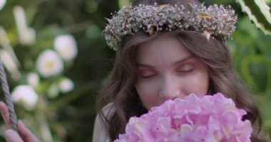 A young beautiful girl in a wreath of flowers swings on a swing among a green garden. Slow motion