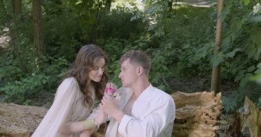 A pair of young lovers in light white robes sit on a log in the garden among flowering plants, holding a rose flower in their hands