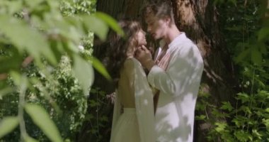 A young pair of lovers in light white robes stand by a large tree in a green flowering garden. Slow motion