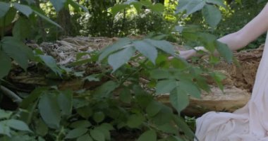 A young woman in a white light dress sits on a log in a green garden among flowering plants. Slow motion