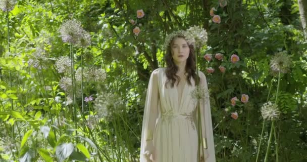 頭に花のリースを持つ若い女性は 緑の葉や開花植物の間で庭の白い光のドレスに立っています スローモーション — ストック動画