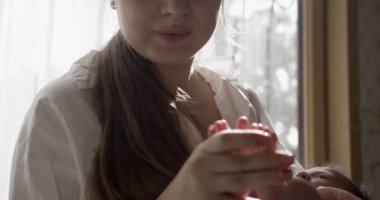 Beyaz tişört giyen anne bebeği elinde tutuyor. - Yakın çekim. Kapalı çekim. Arkasında beyaz perdeler olan pencere.