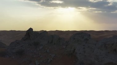 Sızdıran bir kayanın etrafında uçan bir dron, kumlu bir çölün ortasında günbatımı gökyüzünün arka planında duruyor.