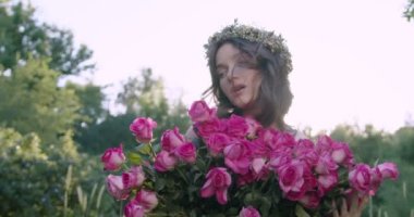A young girl with a wreath of flowers holds a large bouquet of roses among the spring garden. Slow motion