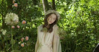 A young woman with a wreath of flowers on her head walks in a white light dress in the garden among green leaves and flowering plants. slow motion