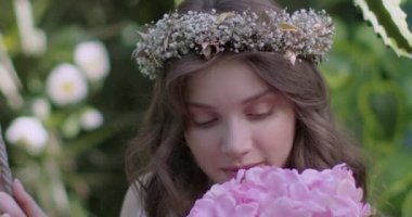 A young beautiful girl in a wreath of flowers swings on a swing among a green garden. Slow motion