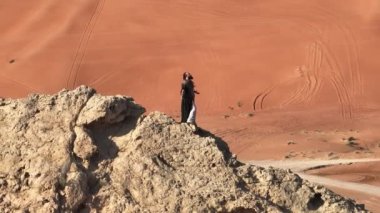Elinde asası ve şaman kıyafetleri olan bir adam çölün kumları arasında bir kayanın üzerinde duruyor ve dans ediyor. Ağır çekim, İHA hava aracı
