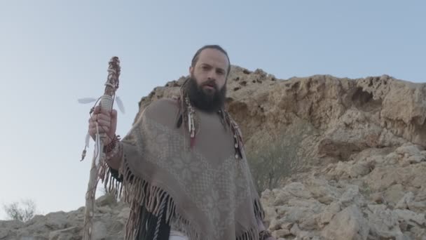 一个留着胡子的年轻人 手拿手杖 披着萨满的衣服 在沙漠的沙丘中的岩石上祈祷 慢动作 阿联酋 — 图库视频影像
