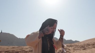 Şaman kıyafetleri içinde genç bir sakallı adam, gün batımına karşı çölde duran siyah bir eşarp. Ağır çekim, BAE