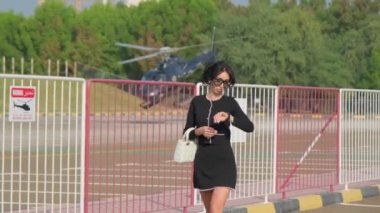 Resmi siyah elbiseli genç bir kadın havaalanından geçen küçük özel bir helikopterin arka planına doğru yürüyor. Yavaş çekim.