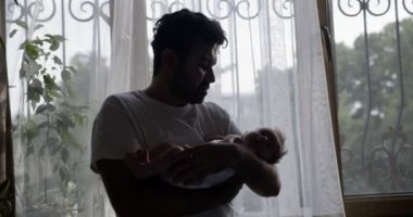 Orta Doğulu adam ellerinde bebek olan beyaz tişörtler giyiyor. İç mekan orta çekim. Arkasında beyaz perdeler olan pencere.