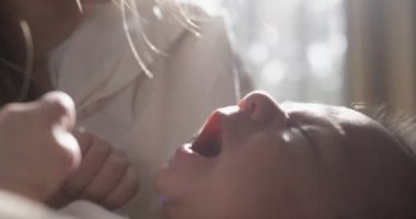 Beyaz tişört giyen anne bebeği elinde tutuyor. - Yakın çekim. Kapalı çekim. Arkasında beyaz perdeler olan pencere.