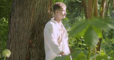 A young man in white light clothes walks past a large tree in a green garden. Slow motion