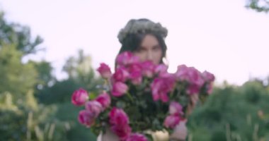 A young girl with a wreath of flowers holds a large bouquet of roses among the spring garden. Slow motion