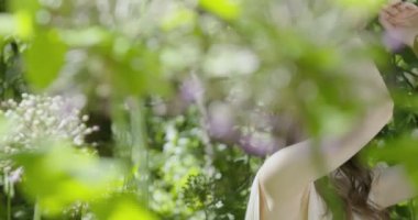 A young woman with a wreath of flowers on her head stands in a white light dress in the garden among green leaves and flowering plants. slow motion