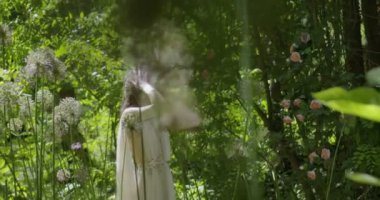 A young woman with a wreath of flowers on her head walks in a white light dress in the garden among green leaves and flowering plants. slow motion