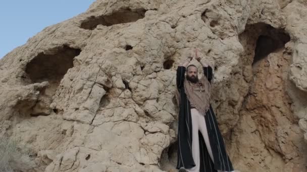 手に杖を持つ男とシャーマンの服が砂漠の砂の間の岩の上に立って踊ります 澄んだ空 スローモーション — ストック動画