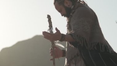 Elinde asası olan genç sakallı bir adam çölün kumlu kumları arasında bir kayanın üzerinde dua ediyor. Ağır çekim, BAE