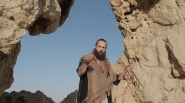 Genç sakallı bir şaman çölün kum tepelerinin arasında bir kaya mağarasında dururken dua eder. Yavaş çekim