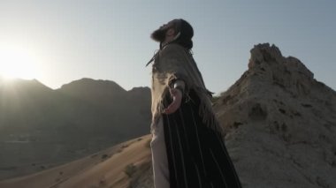 Genç sakallı bir şaman çölün kum tepeleri arasında gün batımının arka planına karşı dua ediyor. Ağır çekim, BAE
