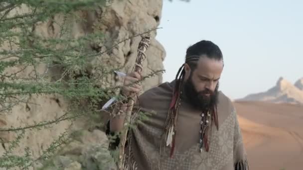 砂漠の真ん中の岩の上を若い髭を生やしたシャーマンが歩いている スローモーション アラブ首長国連邦 — ストック動画