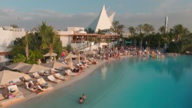 Bir İHA açık havuzun üzerinde, deniz kenarında bir sopayla uçuyor, Dubai. İnsanlar güneşleniyor, barda ve kafede dinleniyor, havuzda yüzüyor. Hava Görünümü.