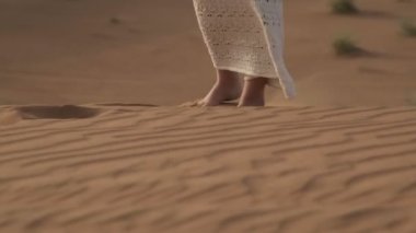 Beyaz elbiseli genç bir kadın çölün kumlarında yalınayak yürüyor.