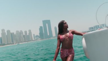 Beyaz bikinili esmer bir kadın ve şehrin arka planına karşı özel bir teknede açık renk bir elbise giyiyor. Ağır çekim, BAE