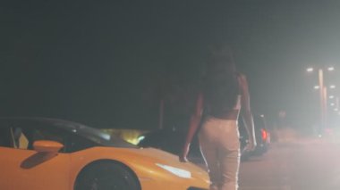 Yüksek topuklu ayakkabı giyen zarif genç bir kadın pahalı bir spor arabaya doğru yürür ve ona biner. Gece, ağır çekim.