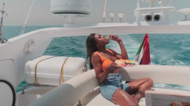 Şortlu ve açık renk bluzlu bir kadın özel bir teknenin arkasında oturur ve elinde tuttuğu bir tabaktan meyve yer. Yavaş çekim.