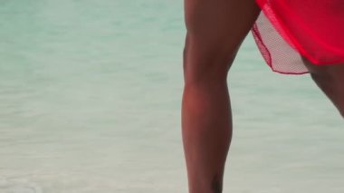 Bikinili esmer ve açık kırmızı elbiseli genç bir kadın sörf suyunda kumlu bir sahilde yalınayak yürüyor. Yavaş çekim