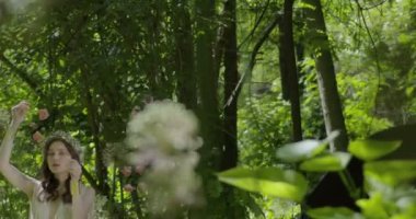 A young woman with a wreath of flowers on her head stands in a white light dress in the garden among green leaves and flowering plants. slow motion