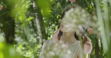 A young woman with a wreath of flowers on her head stands in a white light dress in the garden among green leaves and flowering plants. slow motion