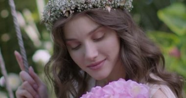 A young beautiful girl in a wreath of flowers swings on a swing among a green garden. Slow motion