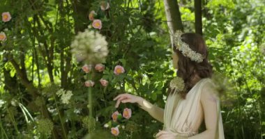 A young woman with a wreath of flowers on her head walks in a white light dress in the garden among green leaves and flowering plants. slow motion