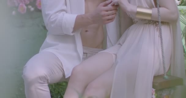 春の庭の中でスイングに軽い白いローブを着た若い恋人たち スローモーション — ストック動画