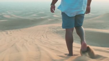 Şort, gömlek ve başörtüsü giyen genç bir adam Birleşik Arap Emirlikleri 'nde çölün kumlarında yalınayak yürüyor. Yavaş çekim