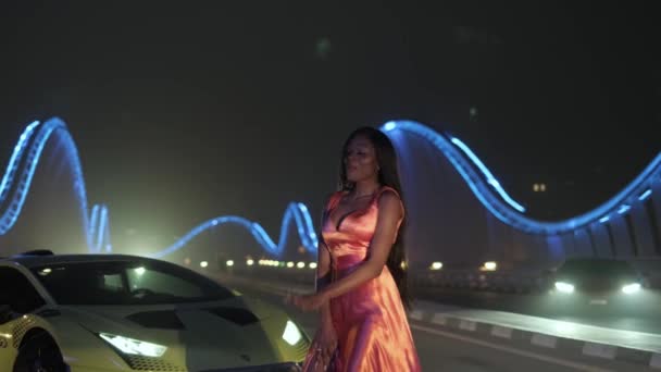 スポーツ車の横にある夜のライトアップされた橋の上でダンスの夜のドレスを着たスワースの若い女性 スローモーション — ストック動画