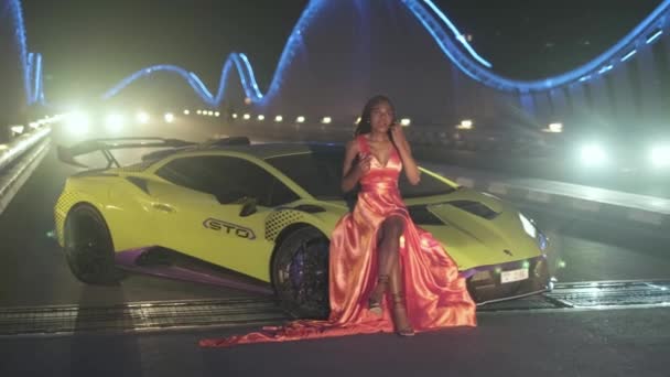 夜にはライトアップされた橋の上にスポーツカーのすすの上に座っている夜のドレスを着た汗ばむ若い女性 スローモーション アラブ首長国連邦 ドバイ — ストック動画