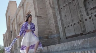 Kısa beyaz elbiseli, uzun saçlı, doğu cüppeli ve kafatası şapkalı genç bir kadın antik bir ahşap kapının önünden geçiyor. Yavaş Hareket, Özbekistan