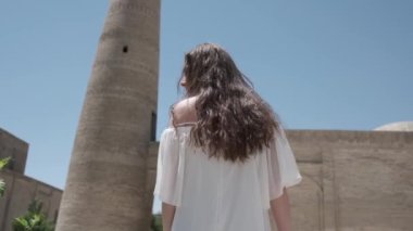 Açık beyaz elbiseli, uzun dalgalı saçlı genç bir kadın antik bir minarenin önünden geçiyor. Yavaş Hareket.
