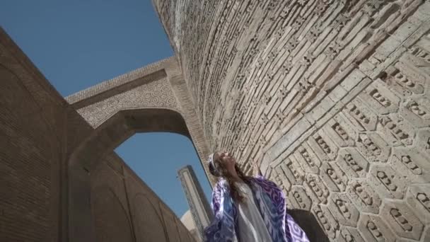流れるような髪の若い女性は 長い東洋のローブと頭皮で ミナレットの前の広場に沿って歩きます スローモーション ウズベキスタン — ストック動画